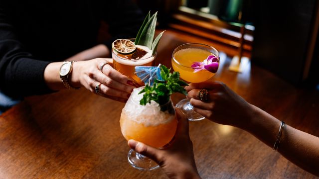 Three people raise colorful cocktails garnished with fruit, flowers, and umbrellas over a wooden table, celebrating together in a cozy, warmly lit setting.