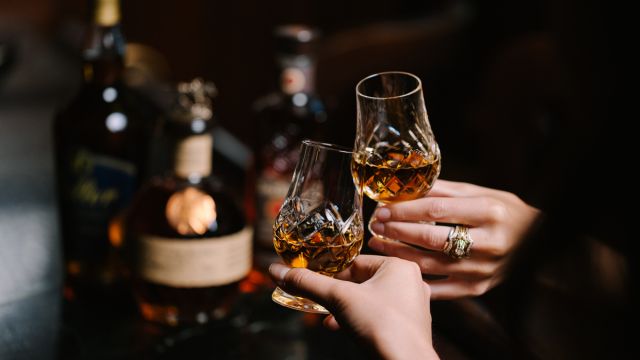 Two hands clinking crystal glasses filled with amber liquor, with several bottles of whiskey blurred in the background on a dark surface.