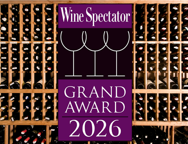A sign reading Wine Spectator Grand Award 2026 with wine glass outlines is displayed in front of shelves filled with wine bottles in a wooden wine cellar.